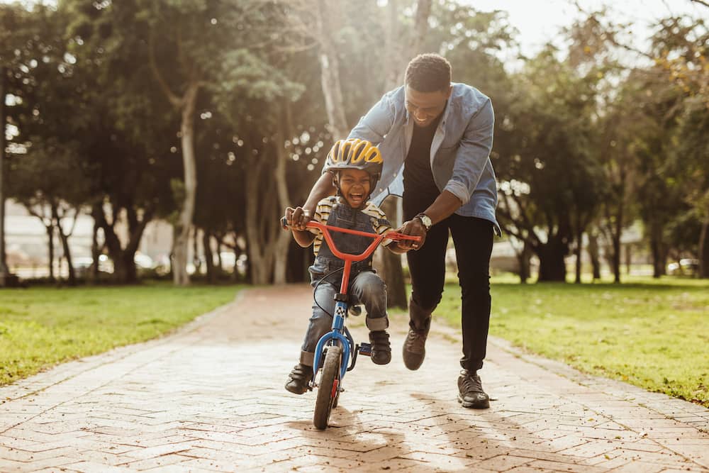 A boy learns to ride his bike with the help of his father in a park.